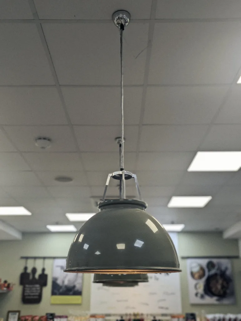 A glossy grey industrial pendant light hanging from a white grid ceiling, with posters and shop displays blurred in the background.