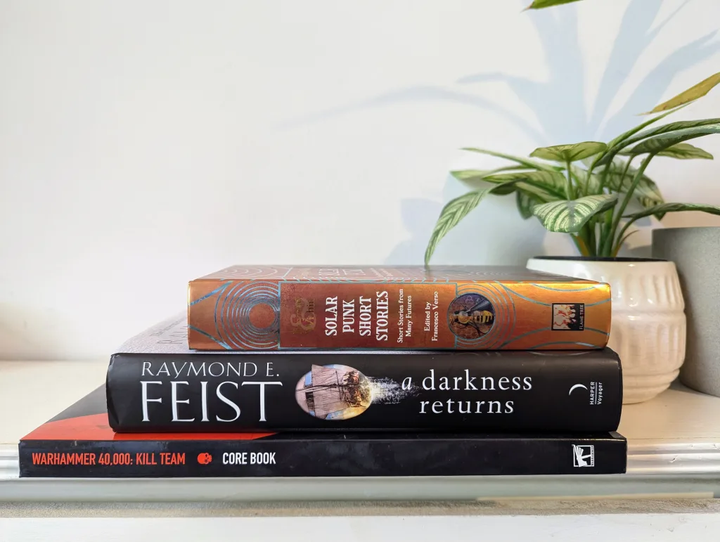 A stack of three books placed on a white shelf next to a potted plant. The books are ‘Solar Punk: Short Stories,’ ‘A Darkness Returns’ by Raymond E. Feist, and the ‘Warhammer 40,000: Kill Team Core Book.
