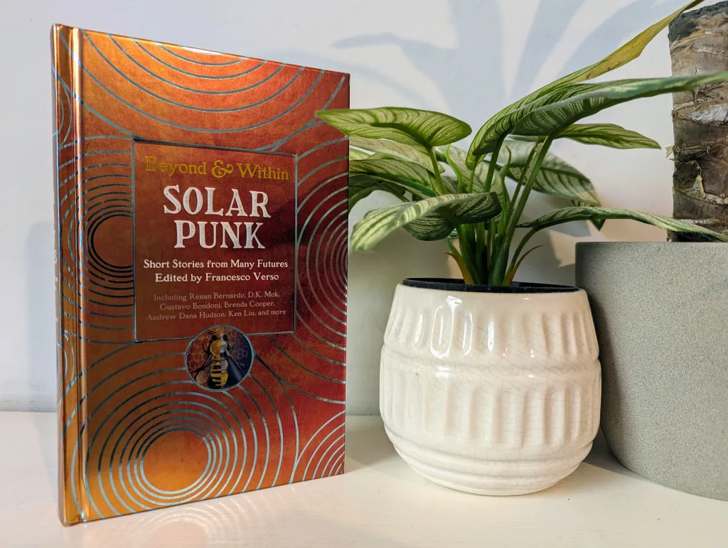 The "Solar Punk" anthology book with a metallic orange cover, standing next to a green variegated plant in a textured white pot.