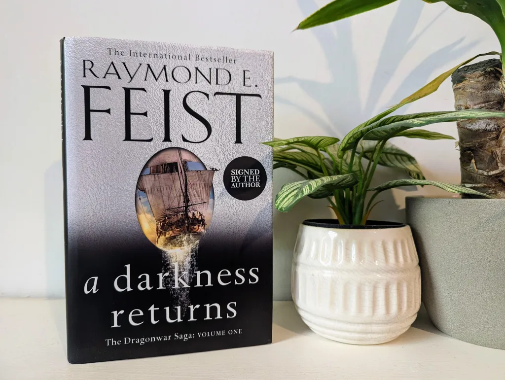 A copy of Raymond E. Feist’s "A Darkness Returns" on a white shelf next to a leafy green plant in a white textured pot.