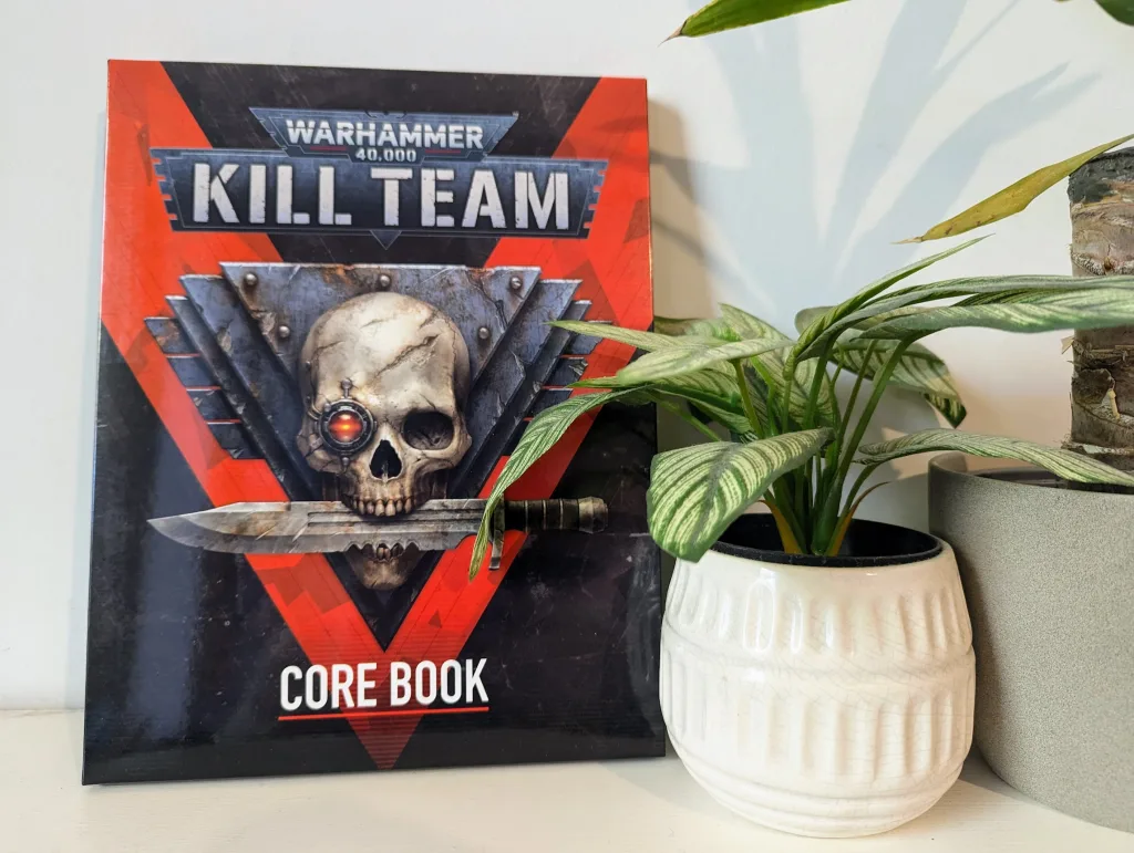 Warhammer 40,000 Kill Team Core Book leaning against a white wall next to a leafy green plant in a white ceramic pot.