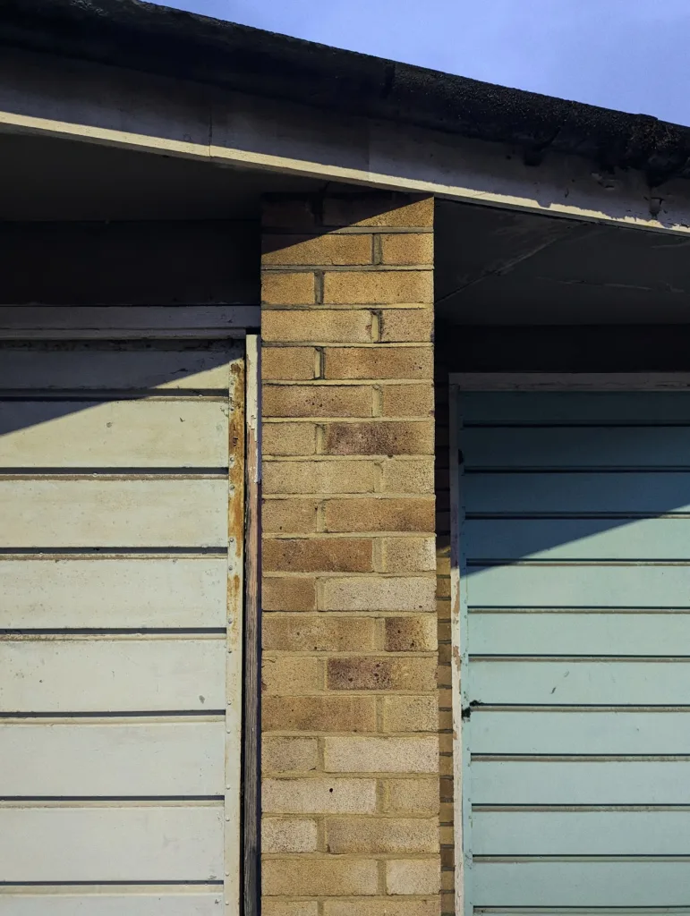 A close-up of a building facade featuring a brick pillar dividing two weathered wooden garage doors, one pale white and the other light blue, with sharp shadows cast by sunlight under a slightly overhanging roof.