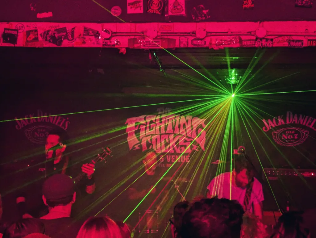 Green laser beams radiate over a band performing on a red-lit stage at The Fighting Cocks venue with crowd silhouettes in the foreground.