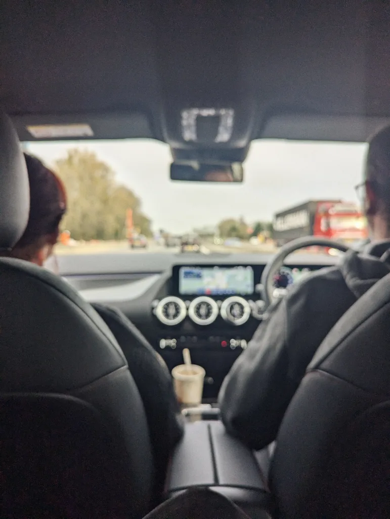 Interior view from the back seat of a car showing two people in front, a dashboard with circular vents, and a red lorry on the road ahead.