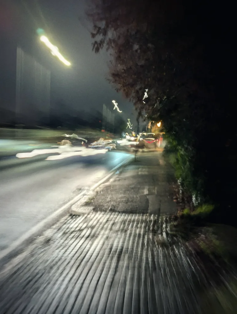Motion-blurred long exposure of a wet city sidewalk at night, featuring streaks of light from traffic against a dark treeline.