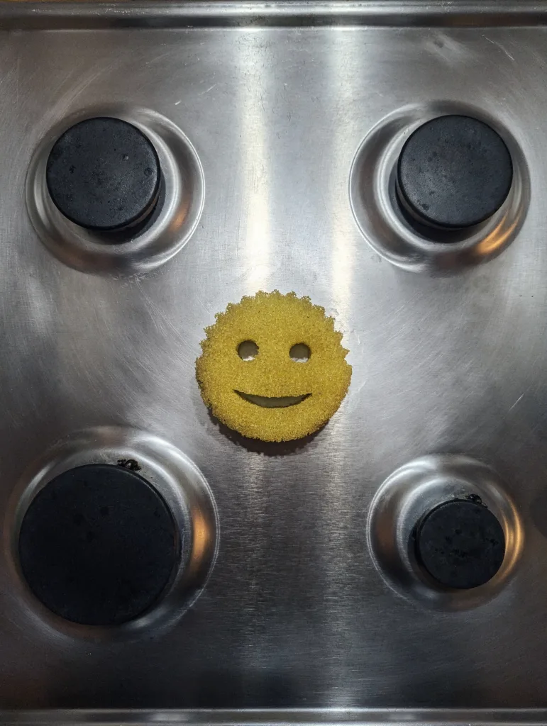 A yellow smiley face sponge centered on a stainless steel stovetop between four black gas burners.