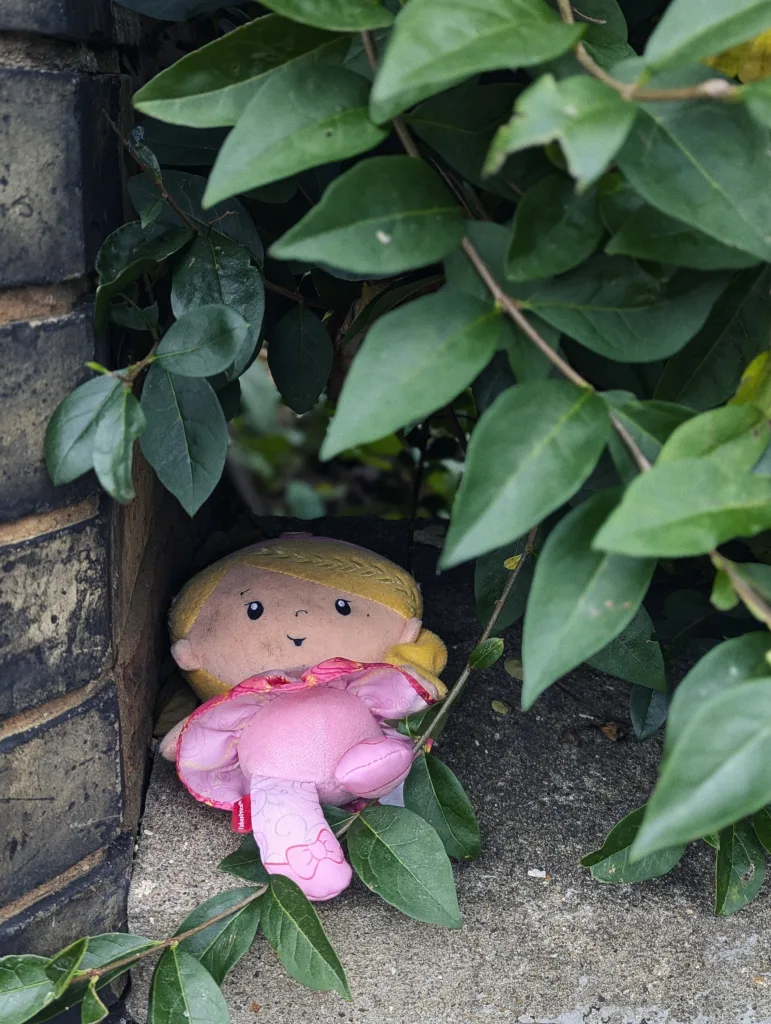 Small blonde plush doll in a pink dress tucked into a concrete nook between a dark brick wall and leafy green branches.