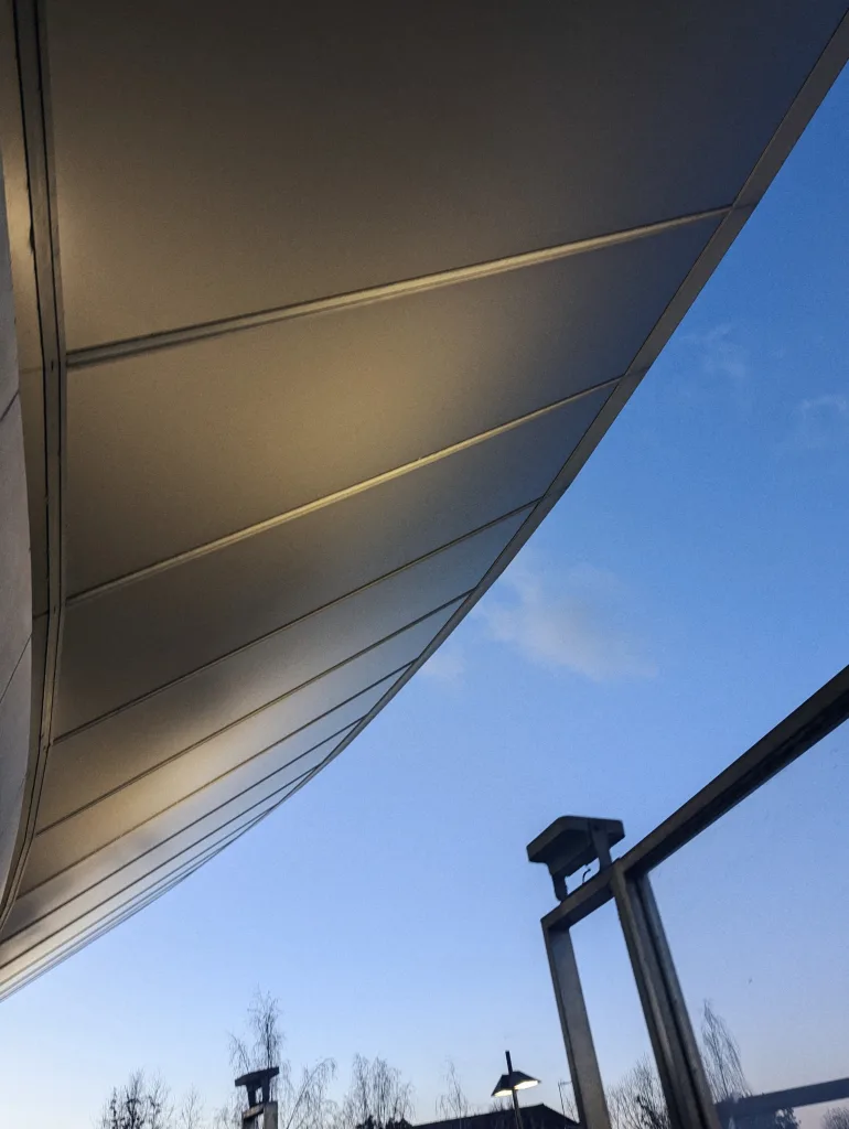 A curved, panelled architectural canopy glows in the warm light of dusk against a clear blue sky.
