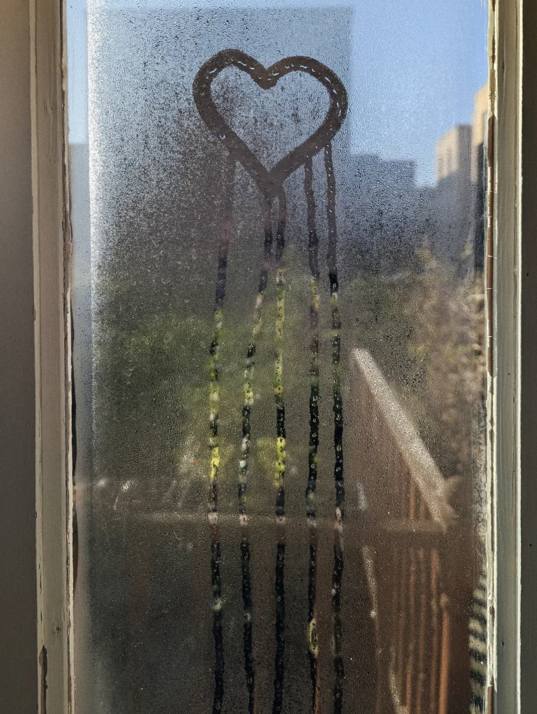A heart drawn in window condensation with four vertical water trails dripping down, overlooking a blurred green garden and blue sky.