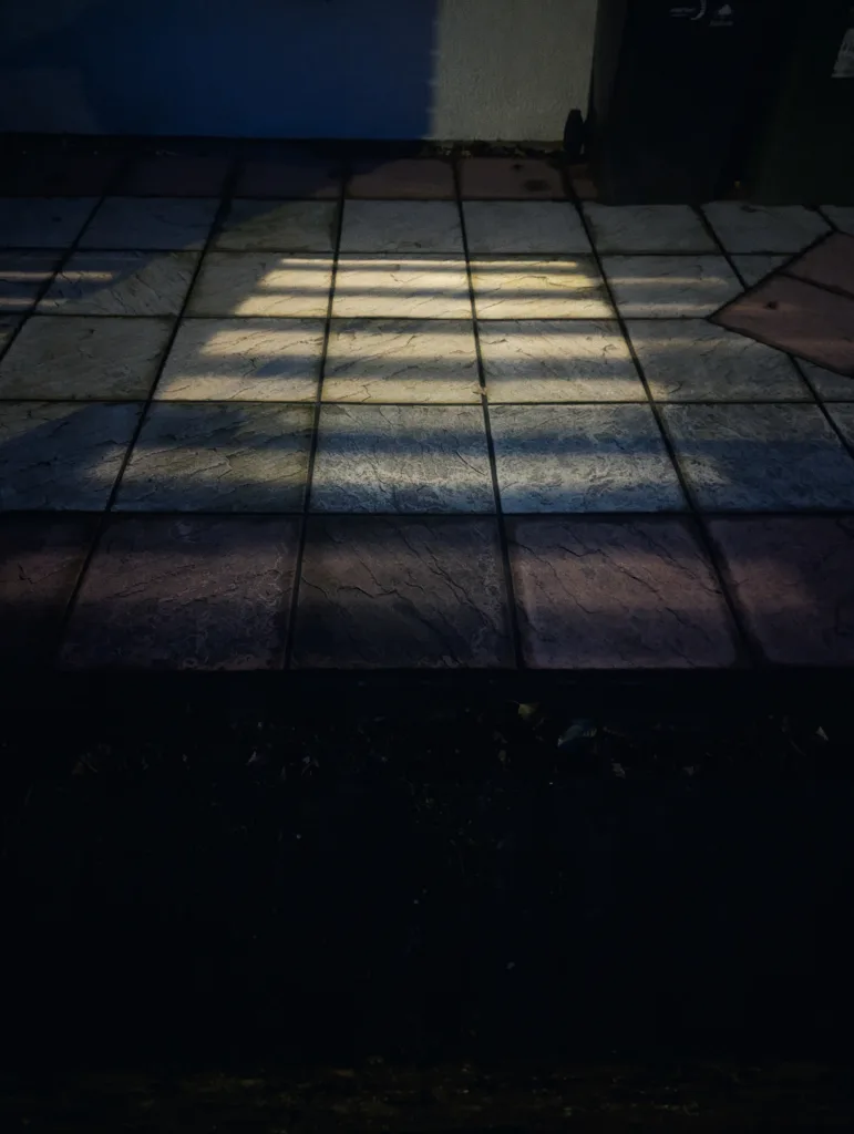 Slatted shadows cast by an unseen fence fall across textured square patio tiles in dim evening light.