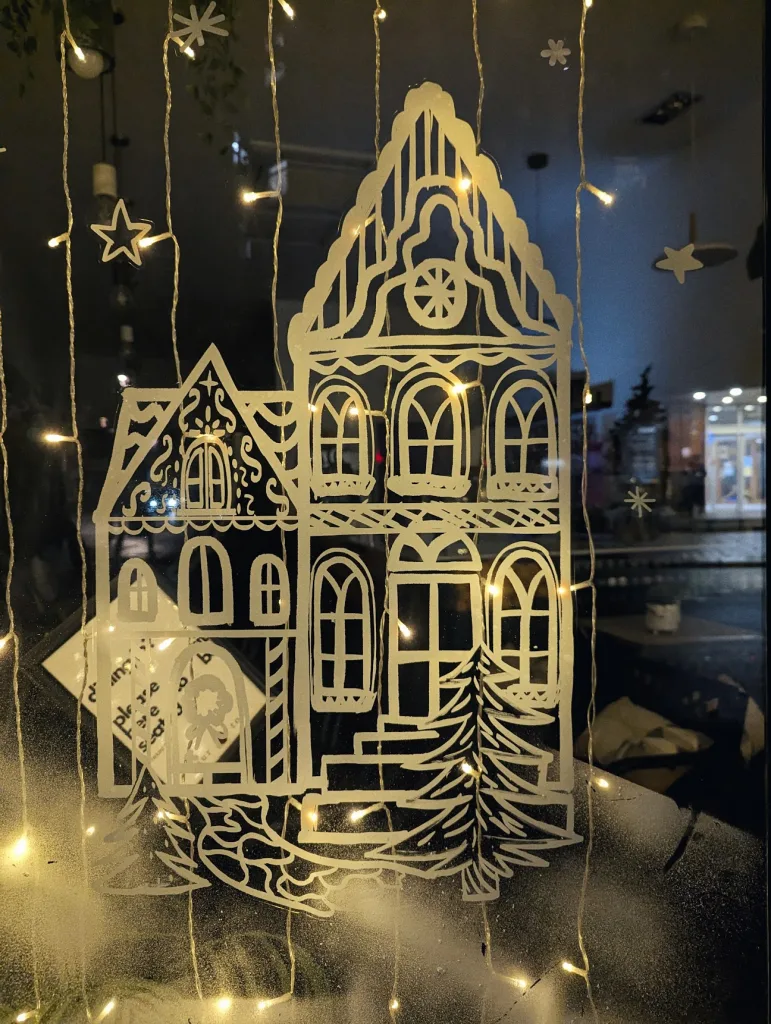 White line art of tall ornate houses on a window, framed by warm hanging fairy lights and snowy spray at the bottom.