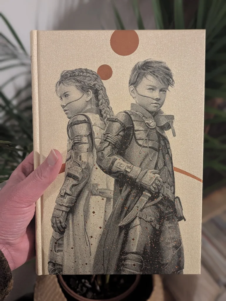A hand holds a gold cloth book cover with a detailed illustration of two young people in desert stillsuits under two orange suns.