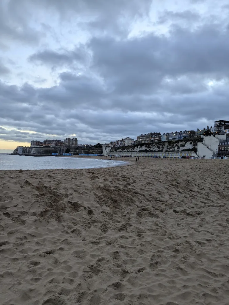 Footprints on a sandy beach leading to chalk cliffs and seaside buildings under a moody grey sky at twilight.