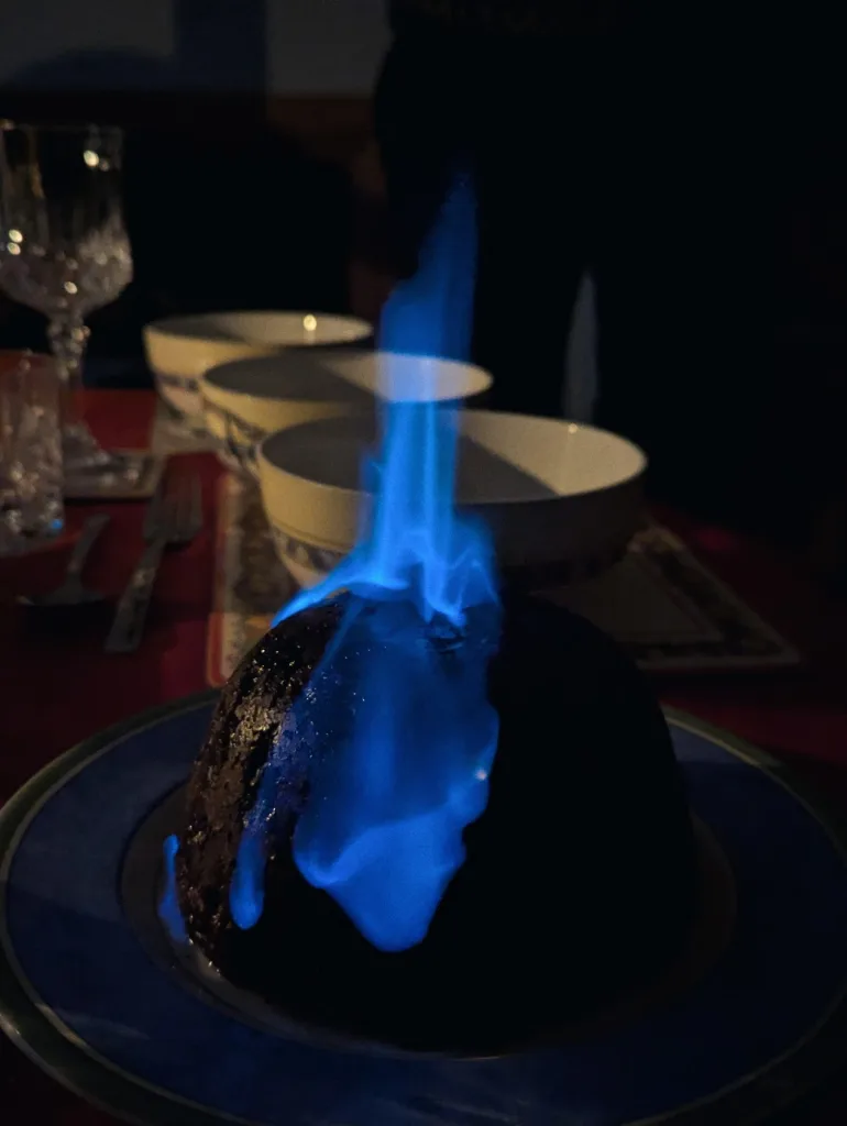 Bright blue brandy flames flickering atop a Christmas pudding on a blue plate, set against a dark dining room background.