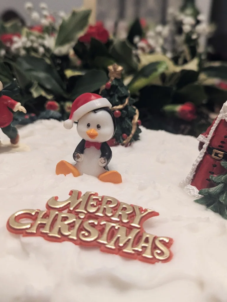 A penguin figurine in a Santa hat on white cake icing with a gold Merry Christmas sign and festive holly in the background.