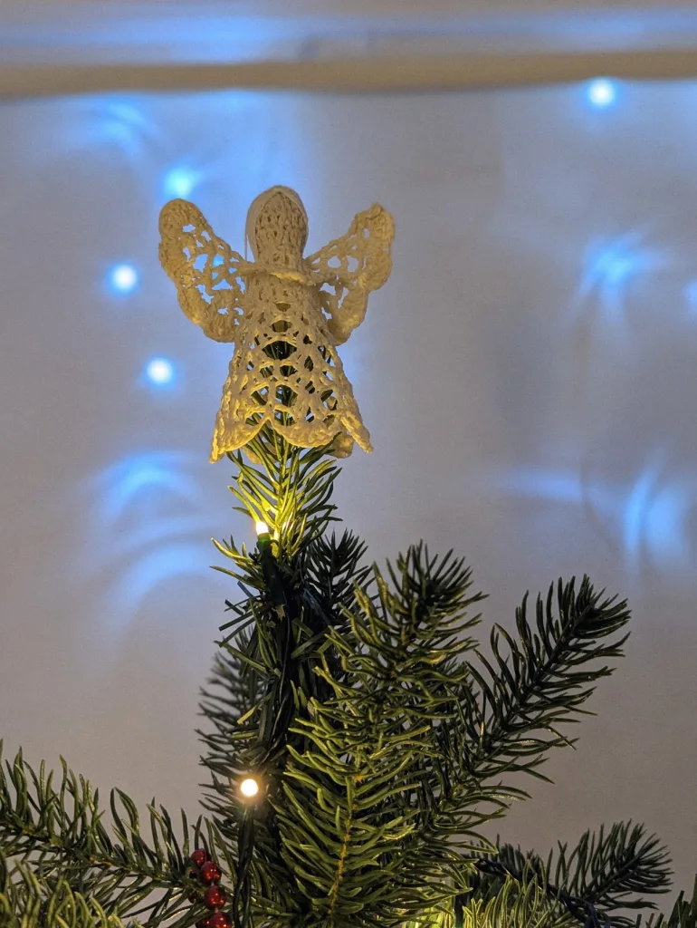 A cream lace angel topper on a Christmas tree, backlit by a warm glow, with blurred blue fairy lights in the background.