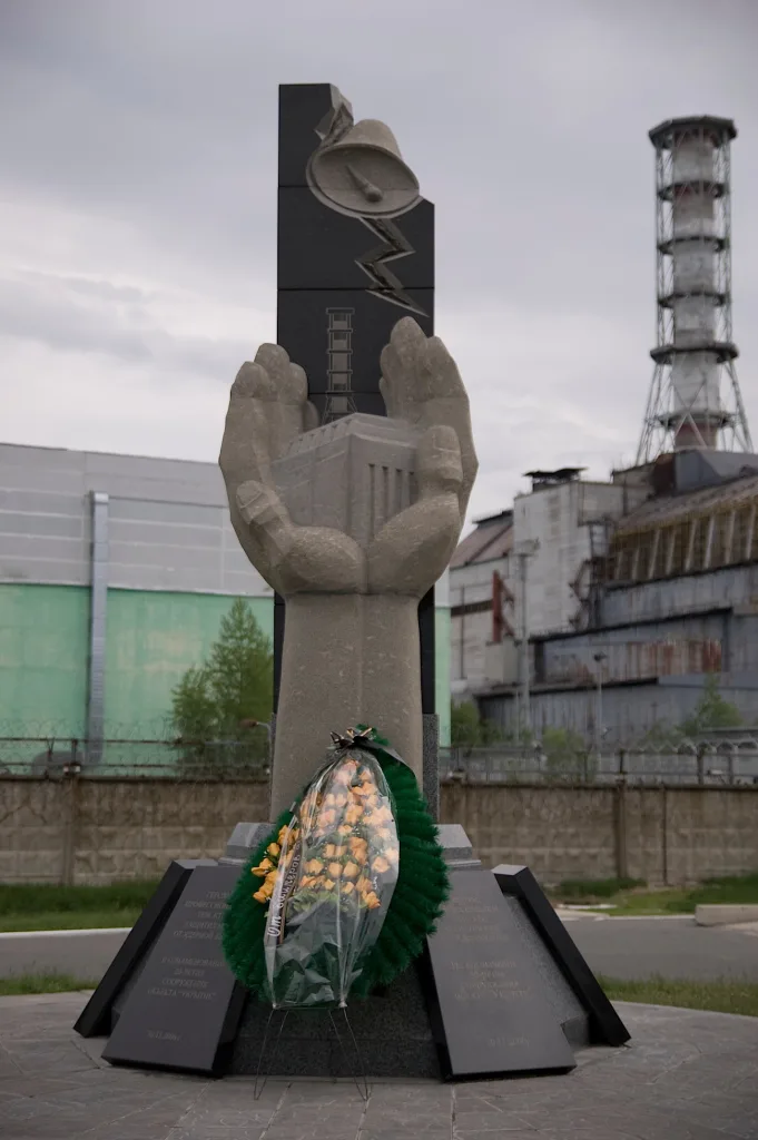 The Chernobyl Liquidators' Monument: stone hands holding the reactor before the power plant and its striped stack under an overcast sky.