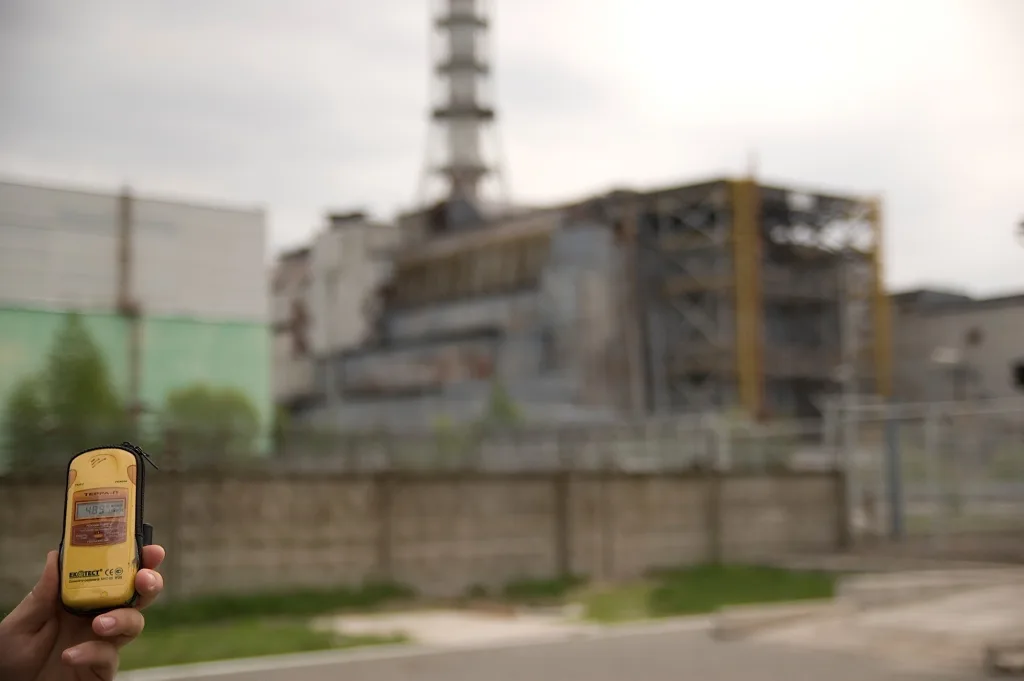 A hand holds a yellow Geiger counter in sharp focus against the blurred backdrop of the Chernobyl Nuclear Power Plant reactor building.