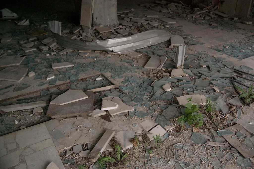 Shattered blue glass shards and broken tiles cover the floor of a derelict building where small green plants are beginning to grow.