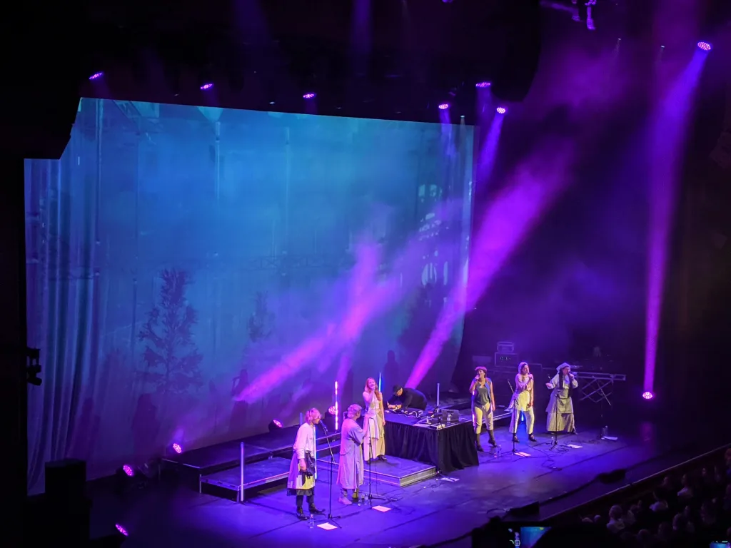 Performers on a stage under purple spotlights, with a large backdrop screen showing a misty forest scene.