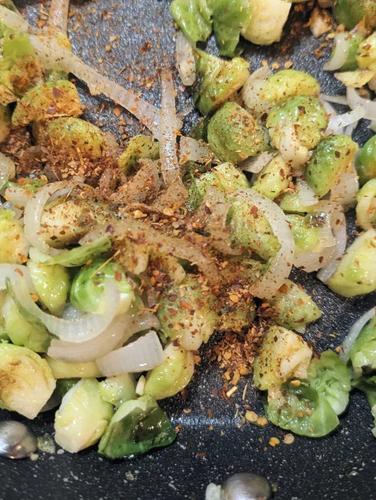 Sautéed brussels sprouts and sliced onions in a dark frying pan, seasoned with red chili flakes and dried spices.