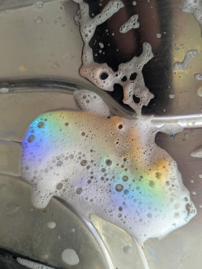 Rainbow light shimmering through a patch of white soap bubbles on a stainless steel sink surface.