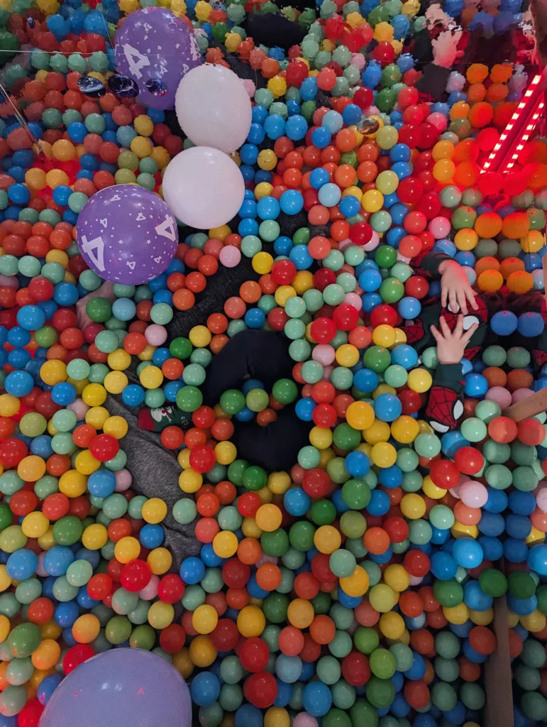 A child's limbs peeking out from a dense sea of multicolored plastic balls, with white and purple number 4 balloons scattered on top.