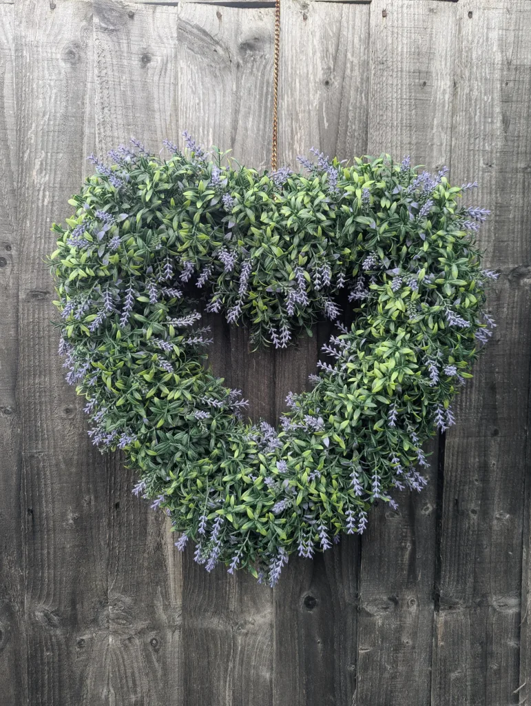 A heart-shaped wreath made of lavender and green foliage hangs from a metal chain against a weathered grey wooden fence.