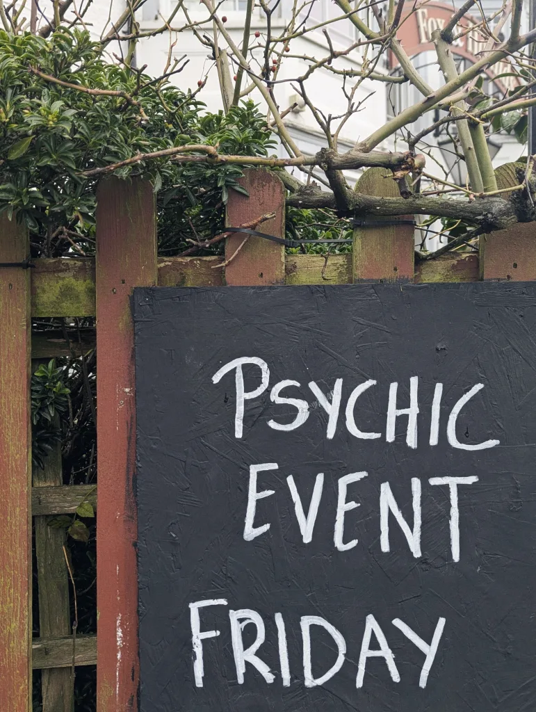 Handwritten white text on a black sign reading "PSYCHIC EVENT FRIDAY" attached to a weathered wooden fence with overhanging greenery.