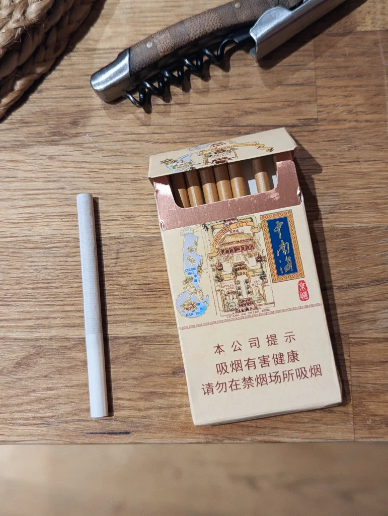 An open pack of Chinese cigarettes featuring a map of the Forbidden City, one loose cigarette, and a corkscrew on a wooden surface.