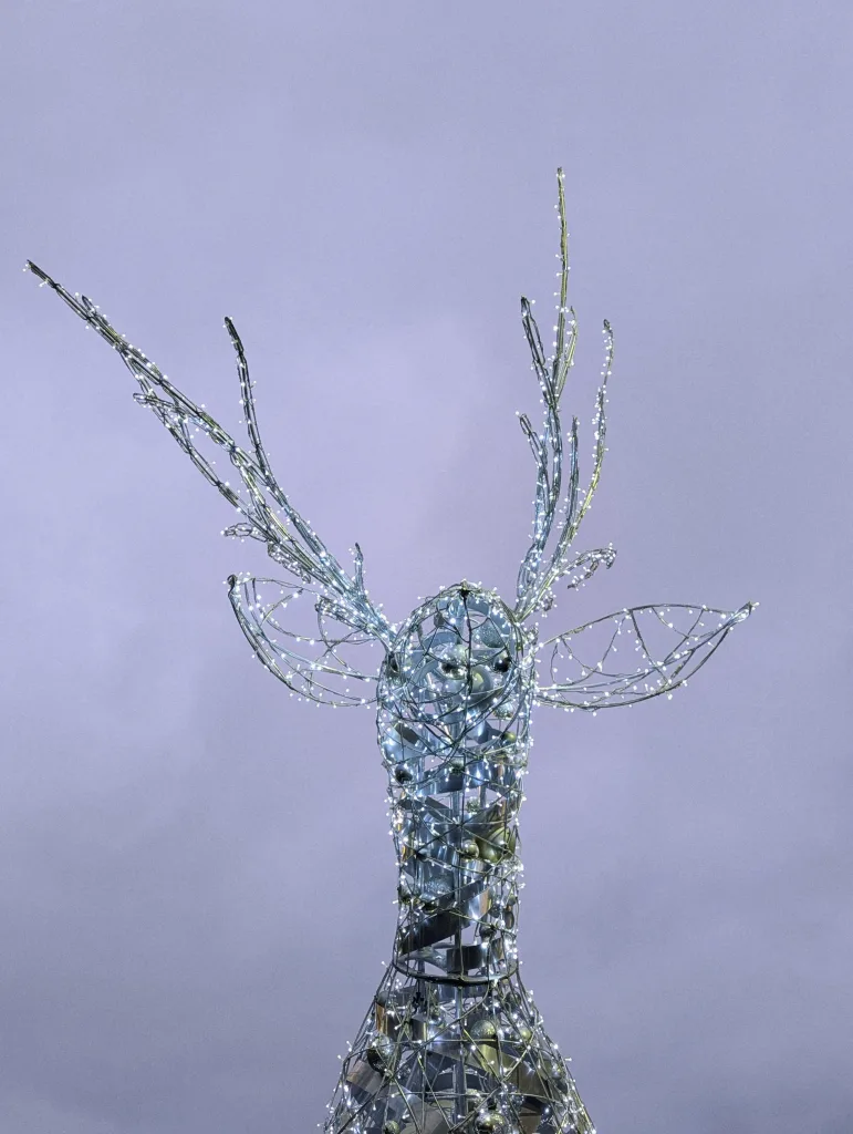 Reindeer head sculpture constructed from silver wire mesh and glowing white lights against a soft lavender background.