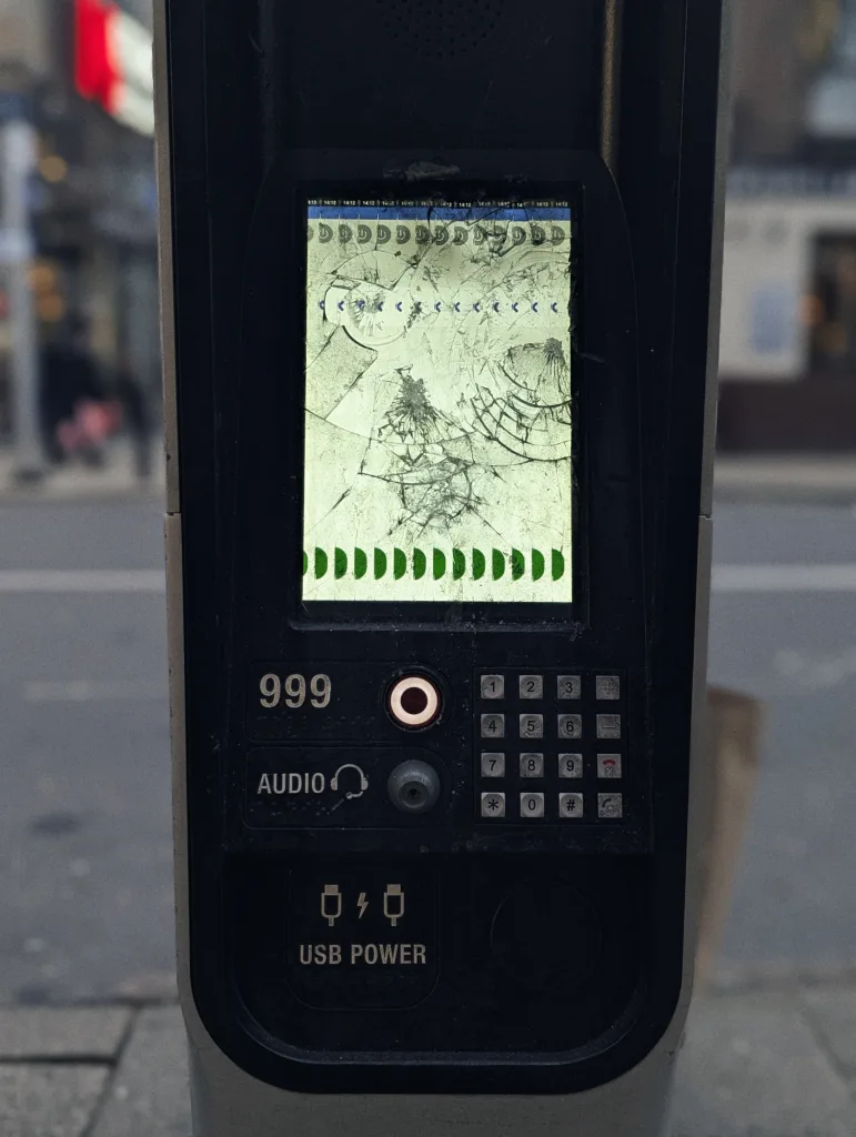 A public street kiosk with a shattered screen displaying glitchy green and blue icons above a keypad and emergency 999 call button.