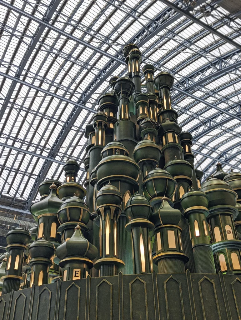 Dark green tiered towers with glowing windows and onion domes, resembling the Emerald City, under a vaulted glass and metal roof.