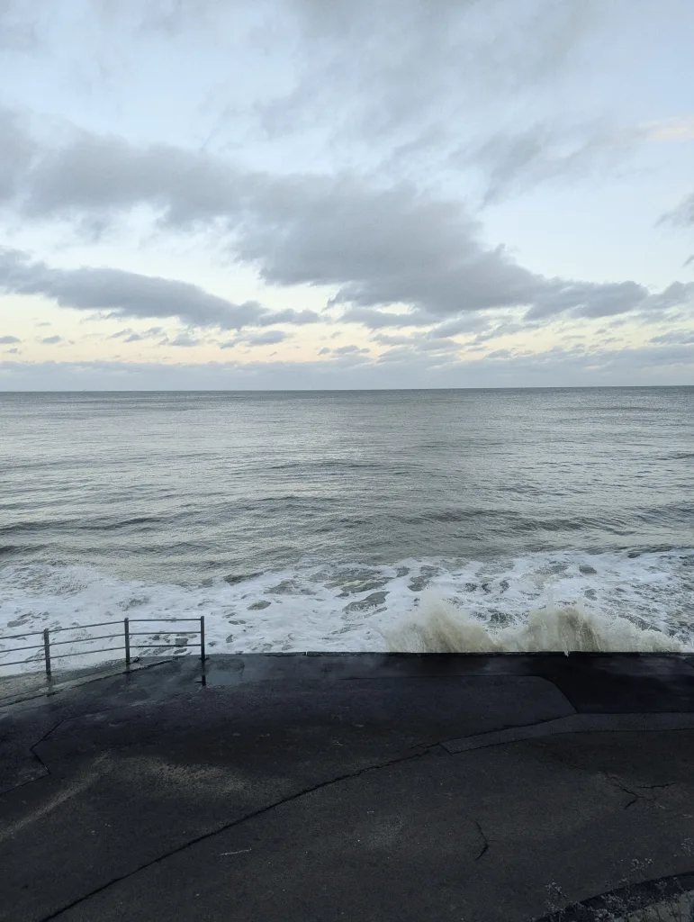 Choppy grey sea waves breaking against a concrete sea wall under an overcast sky.
