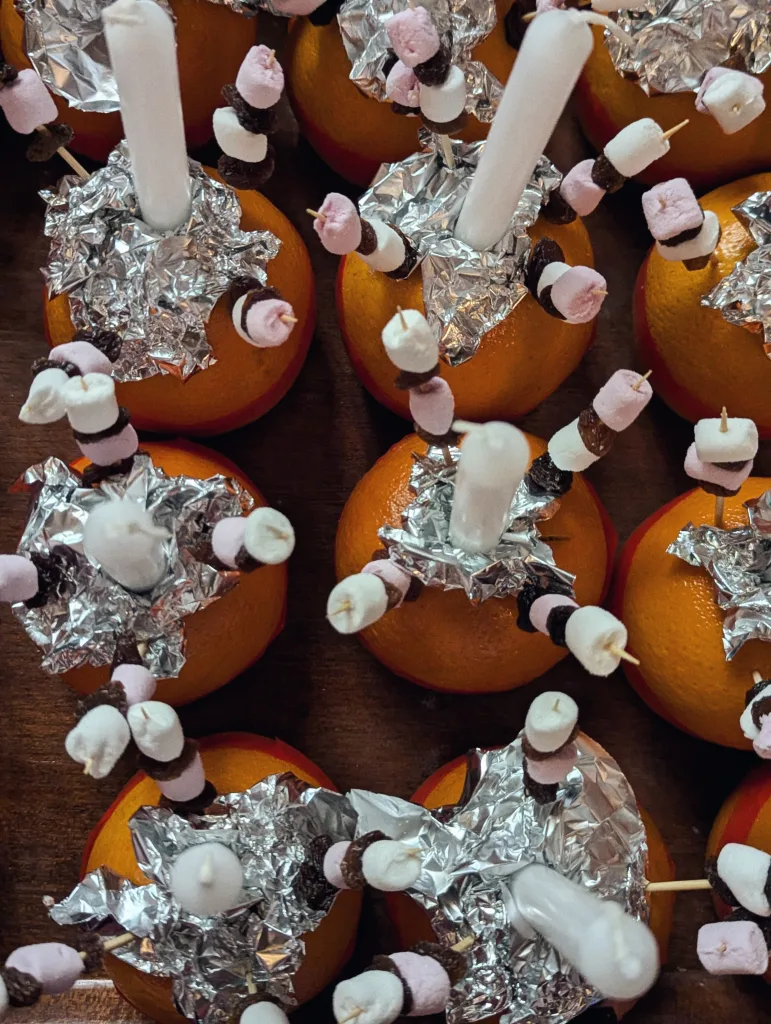 Close-up overhead view of multiple Christingles: oranges with white candles in foil and skewers of marshmallows and raisins.