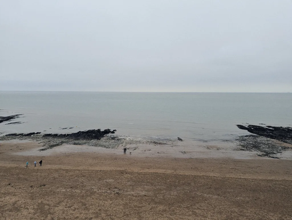 Tiny figures walking on a wide sandy beach between dark rock formations under a pale, overcast sky and calm sea.
