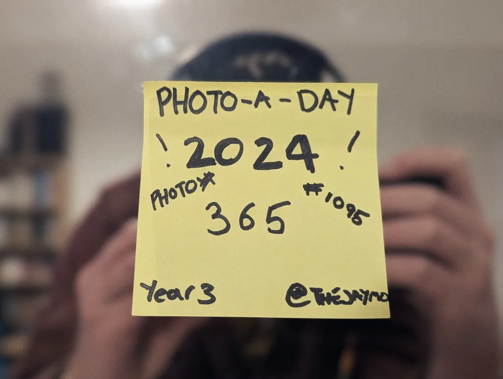 Yellow sticky note on a mirror reading PHOTO-A-DAY 2024 365 #1095 Year 3 @THEJAYMO with a blurry reflection of a person taking a photo.