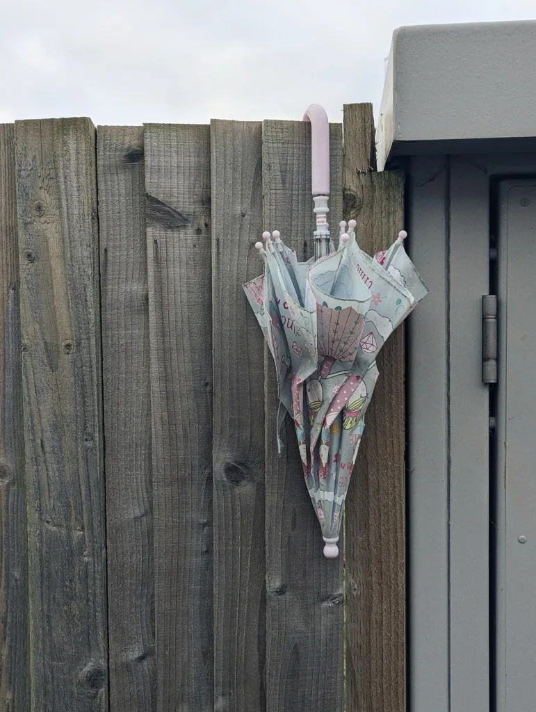 A folded child's umbrella with a pink handle and light blue pattern hanging from a weathered wooden fence panel.