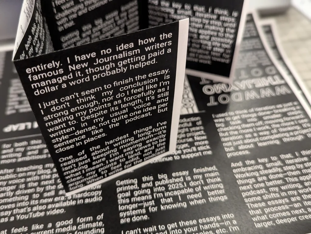 Close-up of a folded black zine with white sans-serif text about writing essays, resting on a sheet with similar printed content.