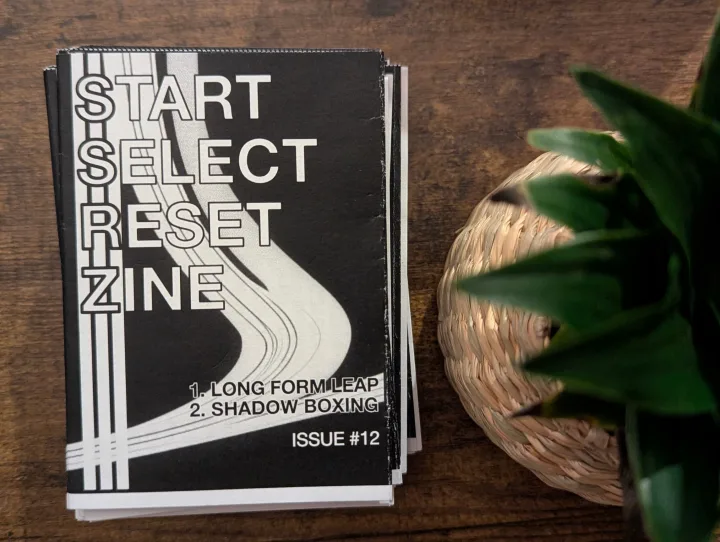 A stack of Start Select Reset Zine Issue 12 on a wooden table beside a small woven basket and green plant leaves.