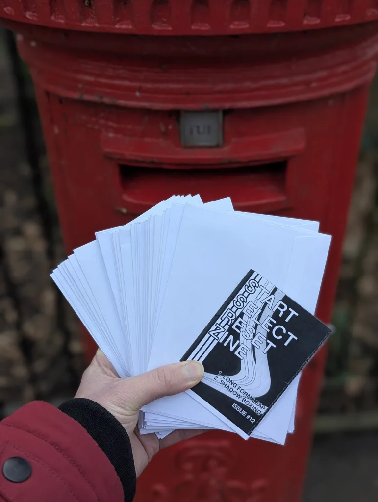 A hand holds a fanned stack of white envelopes and Start Select Reset Zine Issue 12 in front of a classic red pillar post box.