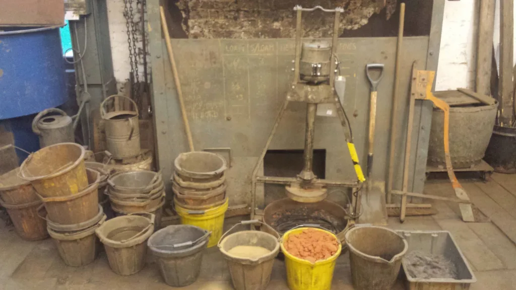 Stacks of buckets and containers of industrial sand and powder in front of a metal loam processing machine in a workshop.
