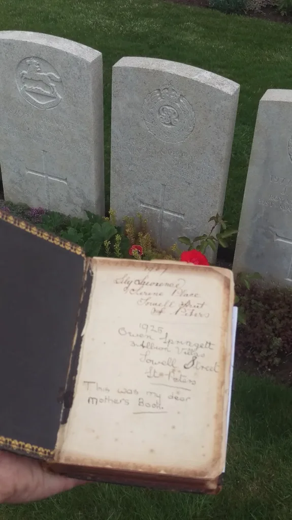 An open vintage book with handwritten family names held in front of the white military headstone for Corporal Henry John Springett.
