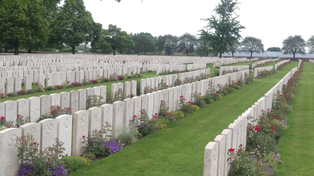 Rows of white military headstones in a green cemetery with vibrant flower beds and leafy trees under an overcast sky.