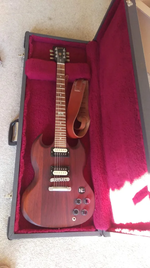 A dark mahogany Gibson SGJ electric guitar with a brown leather strap, resting in an open hard case with a plush magenta interior.