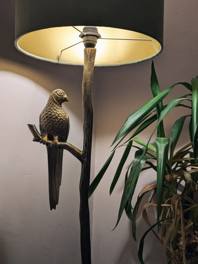 Bronze parrot sculpture on a branch-like floor lamp stand with a dark green shade, next to a leafy houseplant against a pale pink wall.