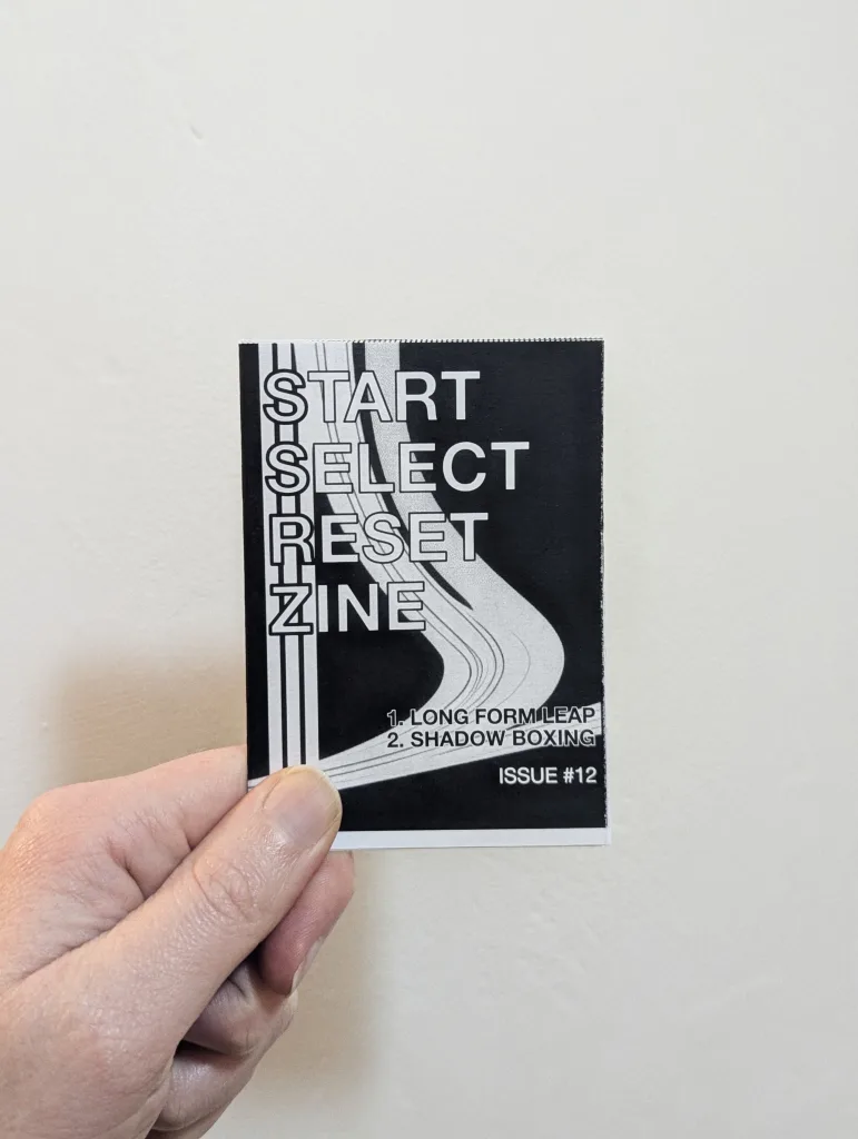 A hand holds the black and white Start Select Reset Zine Issue #12, featuring graphic waves and vertical lettering against a plain wall.