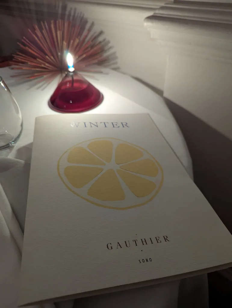 A Gauthier Soho winter menu with a yellow lemon illustration on a white table, beside a lit candle in a red glass holder.
