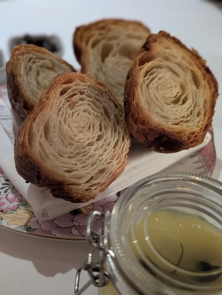 Cross-sections of golden croissants showing intricate flaky layers, served on a floral plate beside a glass jar of yellow curd.