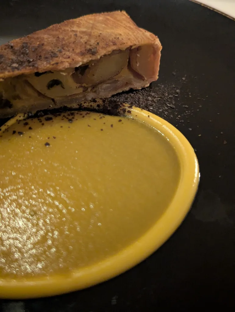 A slice of savory golden pastry served with a smooth circle of yellow puree and dark seasoning on a matte black plate.